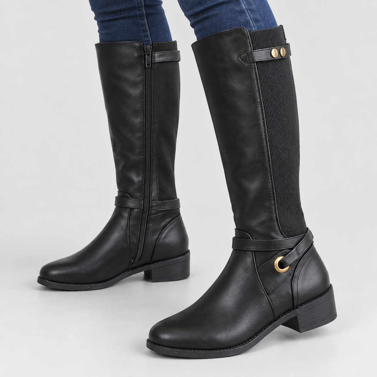 Black tall riding boots with stretch panel and ankle ring worn by a model against a light grey studio background